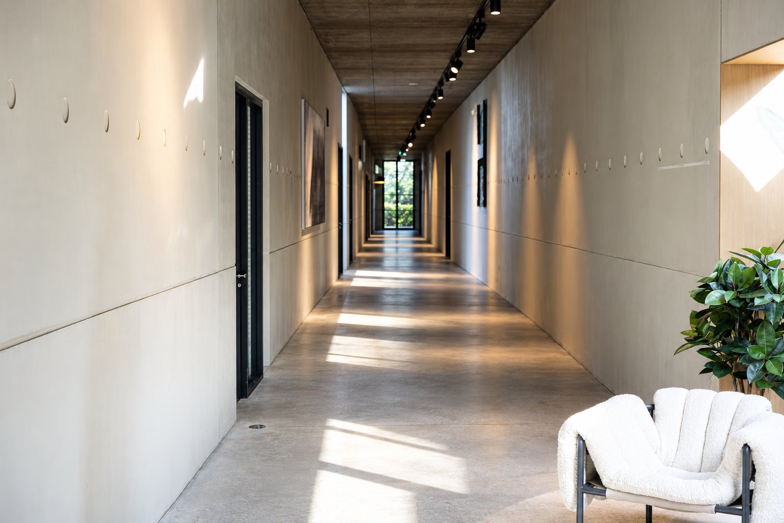 Lichte betonnen gang met plafondspots, kunst aan de wand en een witte bouclé-fauteuil